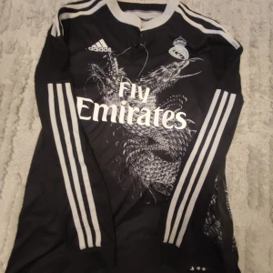 Real Madrid 13/14 Third Shirt - Snygg svart långärmad Real Madrid tröja med vit drakdesign och Fly Emirates tryck. Adidas logga på bröstet och klubbmärke. Namn och nummer 'Sergio Ramos 4' på ryggen. Perfekt för fans!