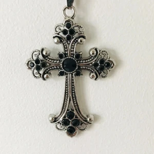 Silverfärgat korshalsband med svarta stenar - Elegant korshalsband i silverfärg med detaljerade svarta stenar. Korset har en vacker design med små runda stenar som ger en stilren och klassisk look. Perfekt för att ge en extra touch till din outfit.