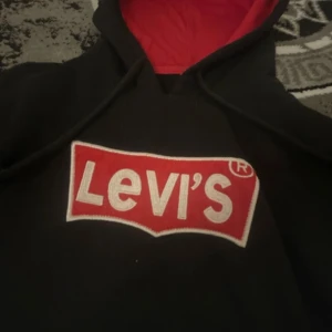 Svart hoodie från Levi's - Säljer en svart hoodie från Levi's med röd insida i huvan och det klassiska Levi's-logotypen på bröstet. Tröjan har långa ärmar och en praktisk magficka framtill. Perfekt för en avslappnad stil.