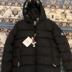 Svart dunjacka från Moncler - Säljer en snygg svart dunjacka från Moncler med huva och dragkedja. Jackan har Moncler-logga på ärmen och är perfekt för kyliga dagar. Den är fylld med vitt gåsdun för extra värme. Kan gå ner i pris mycket vid snabbt köp