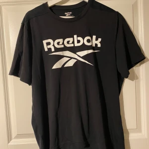 Svart t-shirt från Reebok - Säljer en svart t-shirt från Reebok med stort vitt logotryck på framsidan. T-shirten är kortärmad och tillverkad i ett bekvämt material med Speedwick-teknologi för att hålla dig torr under träning. Perfekt för sportiga aktiviteter eller en avslappnad stil. Sitter som M