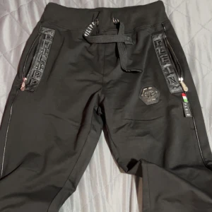 Svarta byxor och kofta från Philipp Plein - Snygga svarta byxor från Philipp Plein med dragkedjor och snörning i midjan. Byxorna har coola detaljer med märkeslogga och en modern design. Perfekta för en trendig look och dem kommer med en kofta som är jätteskön och insidan är jätte snyggt och baksidan av koftan är vacker och den har scan och är aldrig använt