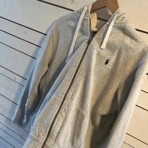 Grå hoodie från Ralph Lauren - Hejsan! Jag säljer en jättefin grå Ralph lauren hoodie med vita snören passar perfekt till våren, den har ett litet hål som inte syns när man bär den i huvan den är väldigt pris värd då den säljs för över 2000kr som ny, inga frågor är dumma!!