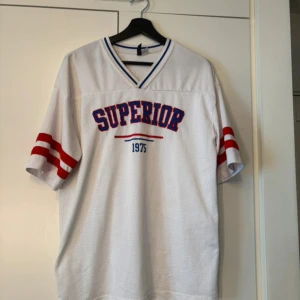 Vit meshtröja med tryck - Säljer en vit meshtröja med texten 'SUPERIOR 1975' i blått och rött på framsidan. Tröjan har en v-ringad hals och korta ärmar med röda ränder. 