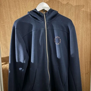 Mörkblå hoodie från Boss - Säljer en stilren mörkblå hoodie från Boss med dragkedja och huva. Perfekt för en avslappnad look. Den har en liten logga på bröstet och långa ärmar. Passar bra till både jeans och joggers.