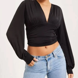Svart omlottopp - Snygg svart omlottopp med långa ärmar och djup v-ringning. Toppen är croppad och har en elegant drapering som ger en smickrande passform. Perfekt för en stilren look. Använd en gång och i mycket bra skick 