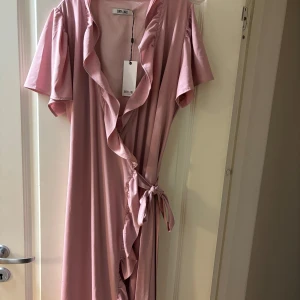 Rosa omlottklänning från Dry Lake - Säljer en romantisk rosa omlottklänning från Dry Lake. Klänningen har korta ärmar och volangdetaljer längs kanten. Perfekt för sommarens festligheter! 💖