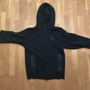 Svart Nike Tech Fleece - Säljer en stilren svart hoodie från Nike med dragkedja och huva. Perfekt för en avslappnad look. Den har långa ärmar och praktiska fickor framtill. Passar bra till både träning och vardag.