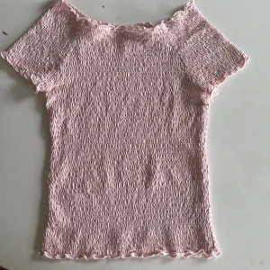 Ljusrosa smockad topp från Lindex - Supersöt ljusrosa topp från Lindex med smockad struktur och korta ärmar. Toppen har volangkant vid hals, ärmslut och nederkant som ger en gullig vibe. Tillverkad i mjuk bomull, perfekt för dig som gillar pastell och comfy stil.