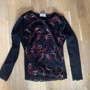 Svart blus med broderade detaljer - Svart blus från Attamis med lång ärm och transparent känsla. Broderade mönster i rött och vitt slingrar sig över hela framsidan. Blusen har en rund halsringning och vågig kant nedtill för extra stilpoäng.