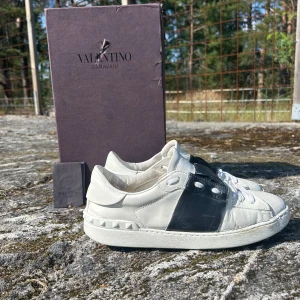 Valentino opens  - Säljer ett par Valentino Garavani Rockstud sneakers i vitt och svart skinn. Skorna har coola nitar runt hälen, vit platt sula och snörning framtill. Ikonisk design med svart panel på sidan och rund tå. Perfekt för dig som vill sticka ut med lyxiga detaljer.