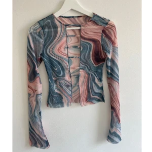 Swirlmönstrad mesh topp XS - Säljer en långärmad mesh topp med swirlmönster i blått, rosa och beige. Toppen har coola cut-outs framtill med smala band över bröstet för en edgy vibe. Materialet är tunn och stretchigt, perfekt för att sticka ut med färg och form.