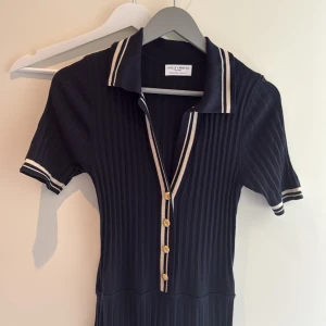 Marinblå långklänning Holly & White - Supersnygg marinblå långklänning från Holly & White med ribbad struktur, korta ärmar och vitrandiga detaljer på krage och ärmslut. Klänningen har guldknappar framtill och en klassisk polokrage som ger en sportig och chic vibe.