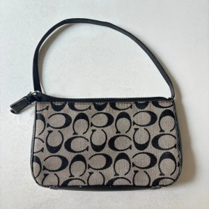 Coach axelväska vintage  - Har tyvärr spilt nagellack på ena sidan, lite sliten längst ner, därav billigare pris. 21x13 cm.            (Snygg axelväska från Coach med klassiskt svart monogrammönster på beige bakgrund. Väskan har en stilren, rektangulär form och dragkedja upptill. Materialet är en mix av bomull och skinn.)
