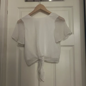 Vit prickig blus med knyt framtill - Säljer en söt vit blus med diskreta prickar och korta, luftiga ärmar. Blusen har en rund halsringning och knyt framtill som ger en chill vibe. Materialet är lätt och känns fräscht mot huden, perfekt för varmare dagar.