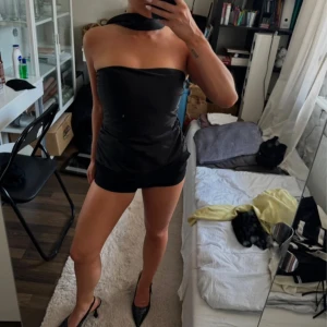 Svart halterneck tubtopp i satin - Supersnygg svart tubtopp med halterneck och lång knytband runt halsen. Toppens material är glansig satin som ger en lyxig känsla. Sitter tight och croppad, perfekt till jeans eller kostymbyxor. Enkel men ändå statement med sin eleganta form.