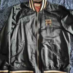 Svart bomberjacka Nudie Jean (M)  - Snygg svart bomberjacka från Nudie Jeans med broderad logga på bröstet och ett stort, coolt kattmotiv på ryggen. Jackan har beige och bruna detaljer på krage och muddar, dragkedja framtill och två fickor. Perfekt för dig som vill sticka ut med streetstyle.
