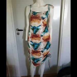 Färgstark kortklänning med knytband - Säljer en cool kortklänning med abstrakt mönster i blått, grönt, brunt och beige. Klänningen har smala axelband med knyt och snygg rynkning längs sidorna. Materialet är mjukt och stretchigt.
