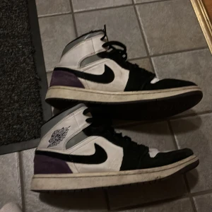 Nike Air Jordan 1 High svart/vit/lila - Nike Air Jordan 1 High sneakers med svart, vit och lila färgblock. Klassisk high-top siluett med snörning, rund tå och platt sula. Ovandel i skinn och mocka, med ikonisk Swoosh på sidan och Air Jordan-logga vid ankeln. Perfekt för dig som vill ha en stilren och sportig look.