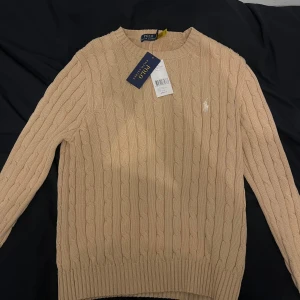 Beige stickad tröja från Polo Ralph Lauren - Snygg beige kabelstickad tröja från Polo Ralph Lauren med rund hals och långärmad passform. Tröjan har det klassiska broderade Polo-logot på bröstet och ribbade muddar vid ärmslut och nederkant. Perfekt för dig som gillar stilren och tidlös design.