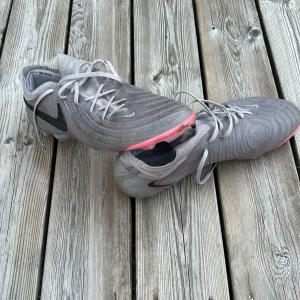 Nike grå fotbollsskor med rosa sula - Säljer ett par grå Nike fotbollsskor med snygg svart swoosh på sidan och ljusgrå snörning. Skorna har en rund tå och en platt sula med coolt rosa och iriserande mönster undertill. Ovandelen är i syntetmaterial som ger bra känsla på planen.
