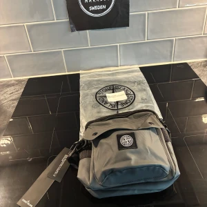 Grå axelväska från Stone Island - Helt ny och oanvänd Snygg grå axelväska från Stone Island med klassisk kompasslogga på framsidan. Väskan har flera fack med dragkedja och tryckknappar, samt justerbar rem. Tillverkad i slitstark polyester som gör väskan vattentät och har coola svarta detaljer. Perfekt för att bära dina essentials.