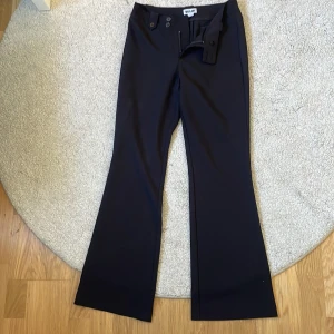 Svarta bootcut byxor från Madlady - Jag är 158 sitter perfekt i längden,Snygga svarta bootcut byxor från Madlady med hög/mid midja och coola knappdetaljer både fram och bak.Användt några få gånger,Byxorna har dragkedja och är gjorda i ett mjukt, slätt material som känns skönt mot huden. Perfekt för dig som gillar en stilren och trendig look.
