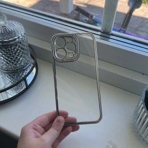 Genomskinlig mobilskal med silverkant - Snygg och stilrent mobilskal till 14 PRO Max med transparent baksida och glittrande silverkant runt hela fodralet. Kameraramen är dekorerad med små stenar för extra bling. Den är använd ett fåtalet gånger och har få tecken på användning.