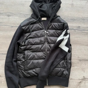 Moncler Cardigan - Svart Cardigan från Moncler med stickade ärmar och huva. Framsidan är quiltad med dragkedja och två fickor. Ena ärmen har en vit zigzag-detalj och Moncler-logga. Snyggt snitt och ribbade muddar för extra stilpoäng.
