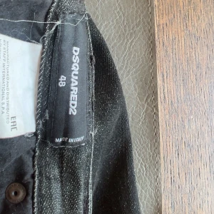 Svarta jeans från Dsquared2, storlek 48 - Snygga svarta jeans från Dsquared2 i storlek 48. Jeansen har en klassisk femficksdesign, slitningar och en cool tvätt som ger dem en edgy vibe. Materialet är jeans med en mörk ton och detaljer som knappgylf och Dsquared2-logga vid linningen.
