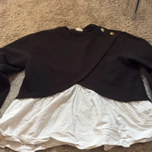 Svart omlott tröja med vita detaljer Zara - Cool svart omlott tröja från Zara med vita volangdetaljer nedtill som ger en lager-på-lager känsla. Guldiga knappar på axeln för extra stilpoäng. Långärmad och perfekt för dig som gillar unika plagg med snygga detaljer.