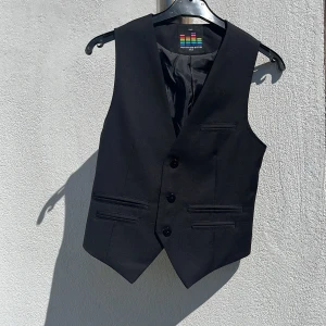 Svart kostymväst Outfitters Nation - Snygg svart kostymväst från Outfitters Nation med klassisk v-ringning och tre knappar framtill. Västen har fyra fickor fram och bröstficka och justerbart bälte bak för perfekt passform. Materialet är slätt och stilrent, perfekt till skjorta och kavaj. Storlek herr XXS och är i mycket fint skick, sparsamt använd och hel o ren. 