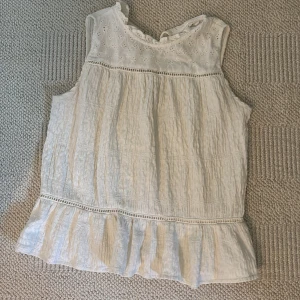 Vit blus med spets från Zara - Supersöt vit blus från Zara med broderad spets upptill och volang längst ner. Blusen är ärmlös och har en lätt skrynklig struktur i tyget, samt dekorativa hålbroderier och små detaljer vid halsen. Den är i storlek 152, men jag skulle säga ungefär xs då den är snygg att som croppad. Priset går att diskuteras