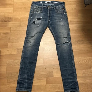 Replay jeans  - Säljer ett par Replay Anbass jeans i slim fit med snygga slitningar och hål för en cool, avslappnad look. Färgen är klassiskt blå med ljusare tvätt och detaljer som bruna läderpatch och fem fickor. Perfekt för dig som gillar streetstyle och vill ha jeans med attityd. 30/32