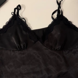 Svart satin linnetopp med spets - Supersnygg svart linnetopp i glansigt satinmaterial med tunna justerbara axelband och spetskant vid bysten. Toppen har en feminin v-ringad form och är perfekt för att ge din outfit en chic touch. Passar dig som gillar stilrena och eleganta detaljer.