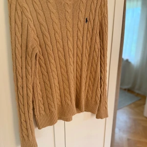 Beige kabelstickad tröja Polo Ralph Lauren - Snygg beige kabelstickad tröja från Polo Ralph Lauren med klassisk rund hals och diskret logga broderad på bröstet. Tröjan har långa ärmar och ribbade muddar vid ärmslut och nederkant. Perfekt för dig som gillar stilren och tidlös design.
