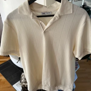 Beige pikétröja från Zara - Snygg beige pikétröja från Zara med diskret ribbad struktur och klassisk krage. Tröjan har korta ärmar och två knappar vid halsen. Perfekt för dig som gillar stilrena och enkla plagg med lite extra känsla i materialet. Pris går alltid att diskutera! 