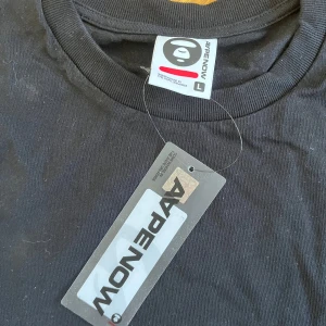 Svart t-shirt från AAPE NOW - Svart t-shirt från AAPE NOW med rund hals och korta ärmar. Diskret broderad AAPE-logga på bröstet och en liten etikett vid nederkanten. T-shirten är gjord i mjuk bomull och har en clean, loose passform som är enkel att matcha med jeans eller shorts.