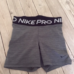 Nike Pro grå träningstights shorts - Nike Pro Dri-Fit shorts i grått med svart elastisk midja och vit logga. Tight passform och midwaist, tillverkade i ett syntetmaterial som andas och transporterar bort fukt. Perfekta för gymmet eller sport, med stilren design och diskret Nike Swoosh på benet.