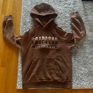 Trapstar hoodie  - Säljer denna bruna hoodie från Trapstar. Tröjan har huva med snörning, magficka och ett  broderat T märke på  luvan. Mjuk insida och ribbade muddar vid ärmslut och nederkant. Passar perfekt för en normal dag.