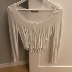 Vit croppad blus med spets från Gina Tricot - Supersöt vit blus från Gina Tricot i croppad modell med lång ärm. Toppen har spetsdetaljer runt halsringningen och framtill, samt en liten rosett. Materialet är mjukt och lätt, och den har en v-ringad design med volangkant nedtill.