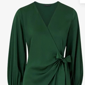 Grön omlottklänning med knyt - Snygg lång grön omlottklänning med v-ringning och knyt i midjan. Klänningen har långa ärmar med knappar vid ärmslut och ett elegant fall. Perfekt för dig som gillar stilrena och trendiga plagg med lite extra detaljer.