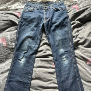 Blå Nudie Jeans - Säljer ett par klassiska blå jeans från Nudie Jeans i straight fit. Jeansen har slitningar i skrevet men som fixats kontrastsömmar. Perfekt passform för dig som gillar en tidlös look. Materialet är robust denim. Hör av er i dm ifall ni har några frågor!😃