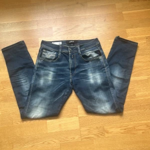 Replay Anbass Hyperflex jeans blå - Snygga Replay Anbass Hyperflex jeans i blå tvätt med slitningar och coola detaljer. Klassisk femficksmodell med raka ben och normal passform. Stretchigt material för extra komfort och rörelsefrihet. Perfekt för dig som gillar en avslappnad men stilren look.