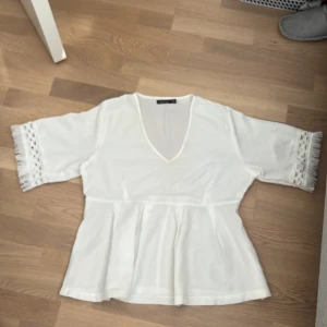 Jättefin top från Boohoo som inte kommer till användning - Säljer en vit blus från Boohoo med v-ringad hals och korta ärmar. Ärmarna har snygga virkade detaljer och fransar som ger en boho-vibe. Blusen har en lätt utsvängd form nedtill och är gjord i ett mjukt polyestertyg. Perfekt för dig som gillar stilrena och trendiga plagg.