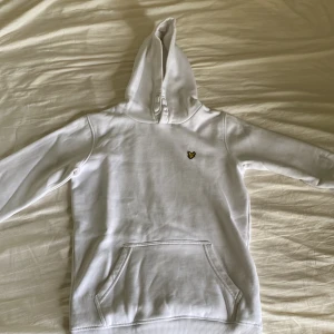 Vit hoodie från Lyle & Scott Junior - Vit hoodie från Lyle & Scott Junior med klassisk gul örn-logga på bröstet. Tröjan har huva, magficka och långa ärmar. Mjuk bomullskvalitet som är skön att ha på sig. Perfekt för chill dagar och enkel att matcha med jeans eller joggers.