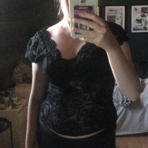 Svart spetsblus med v-ringning - Supersnygg svart blus i spets med v-ringad hals och korta ärmar. Blusen har ett genombrutet mönster och scallop-kant vid halsen. Perfekt för dig som vill ha en feminin och trendig look med lite edge.