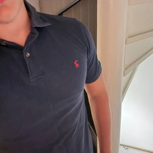 Mörkblå polo t-shirt från Joules - Snygg mörkblå polo t-shirt från Joules i klassisk passform. T-shirten har korta ärmar, krage med två knappar och en röd broderad logga på bröstet. Perfekt för dig som gillar stilrena och enkla plagg med en liten twist.