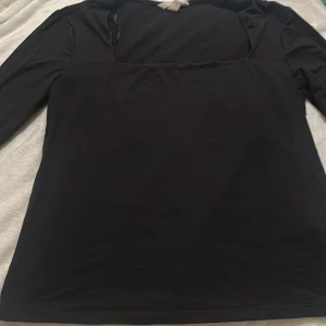 Svart långärmad topp med fyrkantig hals - Snygg svart långärmad topp med en modern fyrkantig halsringning. Materialet känns mjukt och stretchigt, perfekt för dig som gillar stilrena och enkla plagg. Passformen är normal och ärmarna är långa, vilket gör den enkel att matcha med dina favoritjeans eller kjol.