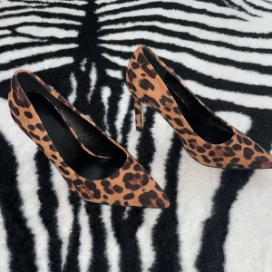 Leopardmönstrade pumps med spetsig tå - Snygga pumps med leopardmönster i bruna och svarta toner. Skorna har spetsig tå och hög klack som ger en riktigt edgy vibe. Utsidan är i mockaliknande material och insidan är svart. Perfekta för dig som vill sticka ut med djurmönster.
